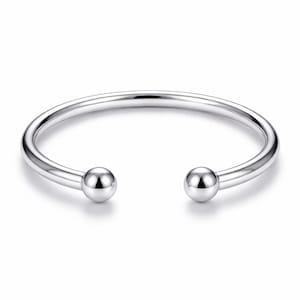 Könnte beinhalten: Ein silberfarbenes, offenes Armband. Das Armband hat ein glattes, abgerundetes Band, das in zwei kugelförmige Endkappen übergeht. Das Armband aus glänzendem, reflektierendem Metall ist vor einem weißen Hintergrund platziert.