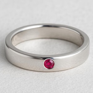 May include: A polished silver ring with a single, round, bright red gemstone. The ring has a flat, smooth band and a minimalist design, showcasing the vibrant ruby. The ring is set against a neutral background.