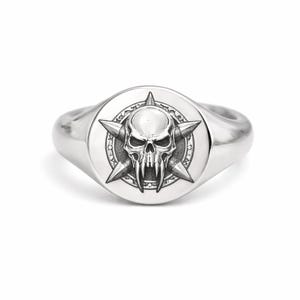 May include: A silver ring with a detailed skull design. The skull is centered on a round face, surrounded by star-shaped points. The ring has a polished finish, reflecting light.