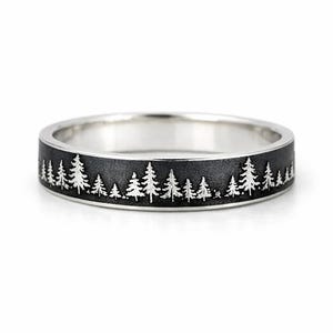 Puede incluir: Un anillo plateado con una banda negra que presenta un diseño de silueta de bosque. El anillo muestra una línea detallada de árboles, creando una estética inspirada en la naturaleza. Es una banda sencilla.