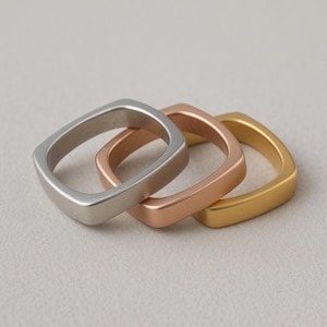 Puede incluir: Tres anillos cuadrados en plata, oro rosa y oro. Los anillos están apilados, mostrando sus diferentes acabados metálicos. Los anillos tienen un diseño moderno y minimalista.