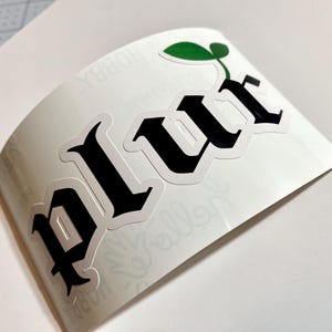 May include: A white sticker with a black gothic font spelling "PLUR" with a green leaf sprout growing from the top of the "R".