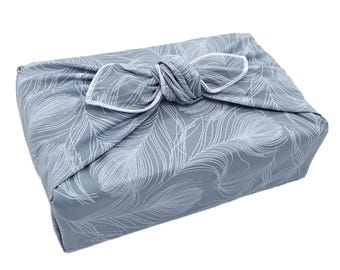 Puede incluir: Envoltorio de tela gris con un diseño de plumas blancas, anudado. Los bordes están acabados con costuras blancas. Este envoltorio reutilizable es una alternativa sostenible al papel de regalo.