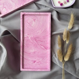 May include: Pink marbled resin trays in rectangular and square shapes. A heart-shaped candle with a lavender rim and small pink flowers sits nearby. Dried wheat stalks add a natural accent to the composition. The trays have a swirled, fluid design.