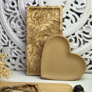 Könnte beinhalten: Ein rechteckiges Tablett mit braunem und cremefarbenem Marmormuster steht neben einem herzförmigen Tablett. Das herzförmige Tablett hat eine einfarbige beige Farbe. Im Hintergrund befindet sich eine dekorative, weiß geschnitzte Tafel.