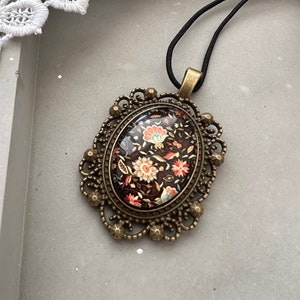 May include: A black cord necklace with a bronze oval pendant featuring a floral design in shades of red, orange, and white on a black background.
