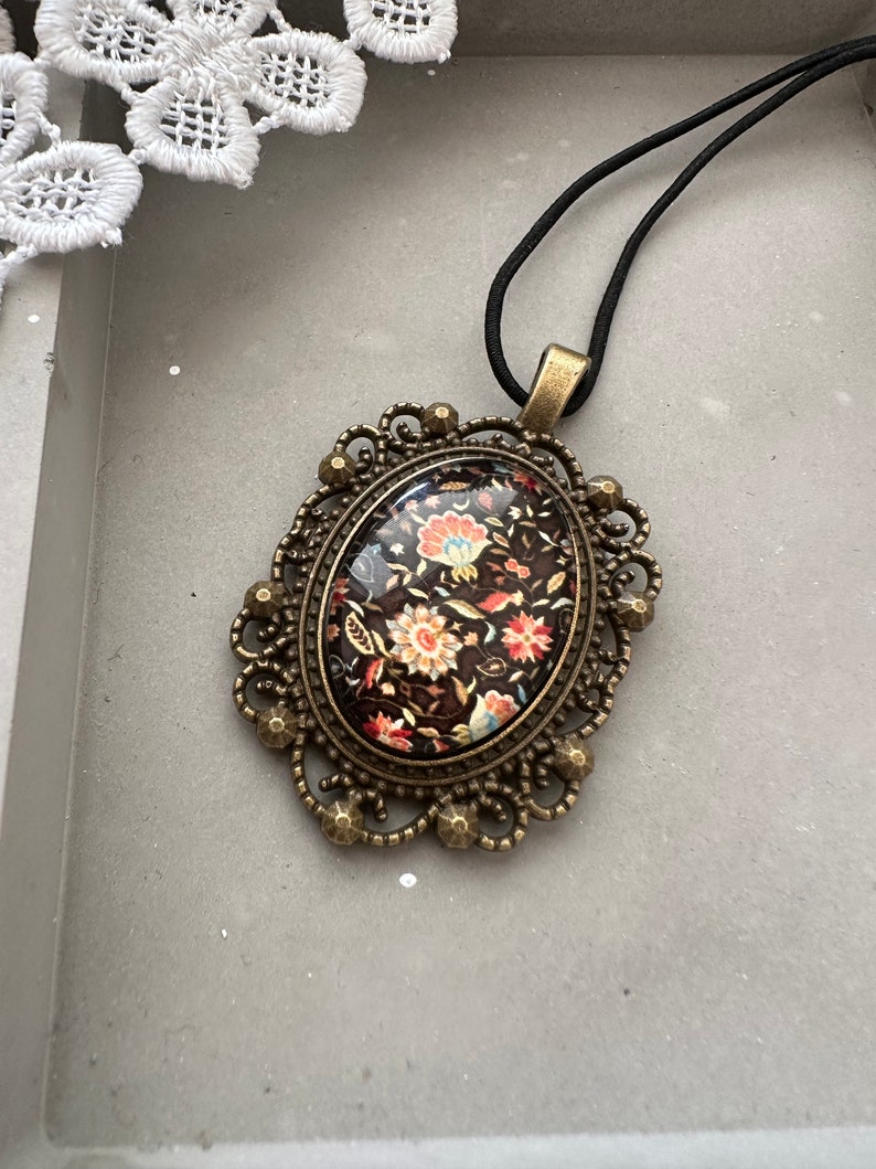 May include: A black cord necklace with a bronze oval pendant featuring a floral design in shades of red, orange, and white on a black background.