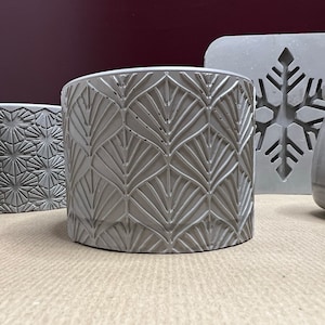 Puede incluir: Cuatro macetas de hormigón gris con diferentes patrones en relieve. Las macetas están sobre una superficie marrón claro. Una maceta tiene un diseño de copo de nieve.