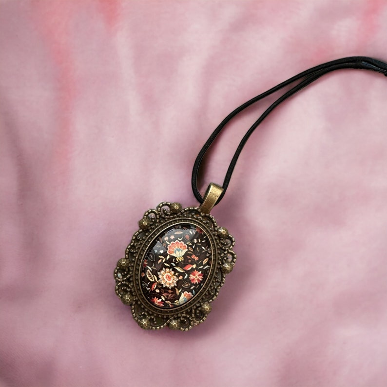 May include: A black cord necklace with a gold oval pendant featuring a floral design in shades of red, orange, and black.