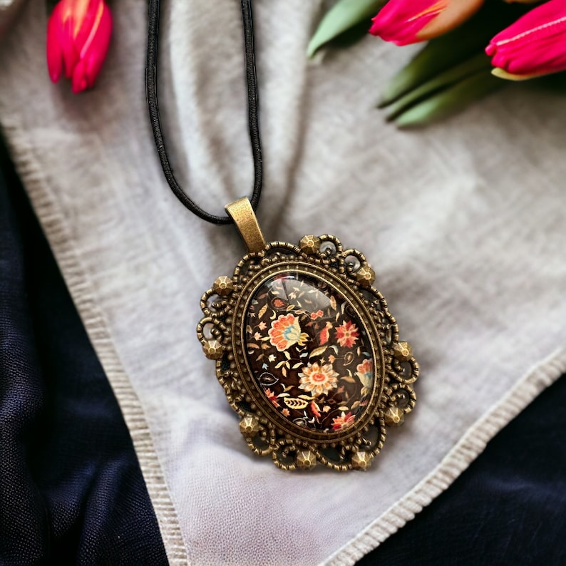May include: A black cord necklace with a bronze oval pendant featuring a floral design in shades of red, orange, and yellow on a black background.