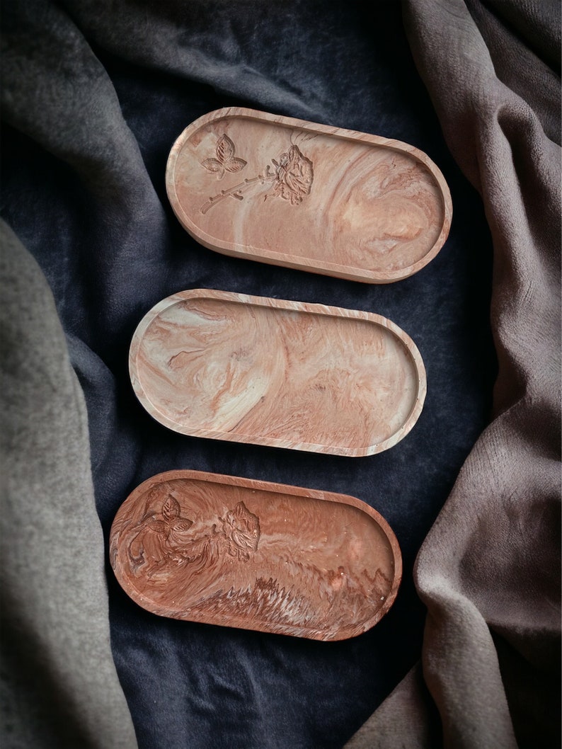 K&ouml;nnte beinhalten: Drei ovale Dekoschalen mit Marmoreffekt in Rosa, Wei&szlig; und Braun. Zwei Schalen weisen ein gepr&auml;gtes Blumendesign auf. Die Schalen sind auf einer dunkelblauen Oberfl&auml;che mit drapiertem Stoff angeordnet.
