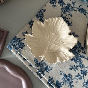 Può includere: Un piatto in ceramica bianca a forma di foglia con un motivo di venature in rilievo. Il piatto è su un tessuto floreale blu e bianco.