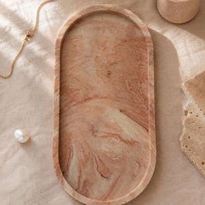 K&ouml;nnte beinhalten: Eine ovale dekorative Schale mit einem marmorierten Muster in Pfirsich- und Cremet&ouml;nen. Eine goldene Kette, eine Perle und eine brennende Kerze in einem kleinen beigen Halter liegen neben der Schale. Die Schale ist ein stilvolles Wohnaccessoire.