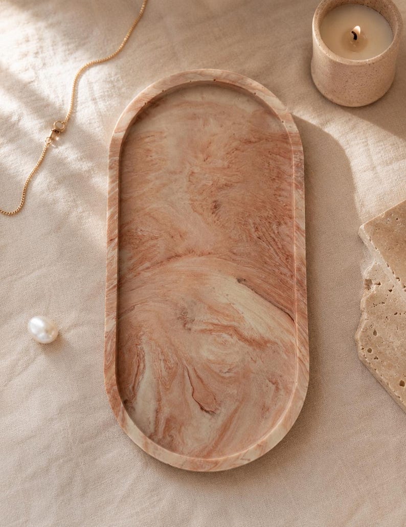 K&ouml;nnte beinhalten: Eine ovale dekorative Schale mit einem marmorierten Muster in Pfirsich- und Cremet&ouml;nen. Eine goldene Kette, eine Perle und eine brennende Kerze in einem kleinen beigen Halter liegen neben der Schale. Die Schale ist ein stilvolles Wohnaccessoire.