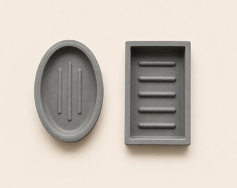 May include: Two grey soap dishes. One is oval with three vertical ridges, the other is rectangular with five horizontal ridges. Both are made of a matte material and sit on a light beige surface. The soap dishes are designed to hold bars of soap.
