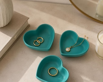 May include: Three teal heart-shaped trinket dishes. Each dish holds jewelry: two gold hoop earrings, a gold necklace with a pearl pendant, and a gold ring. The dishes are arranged on a light-colored surface, with a mirror and candle in the background.