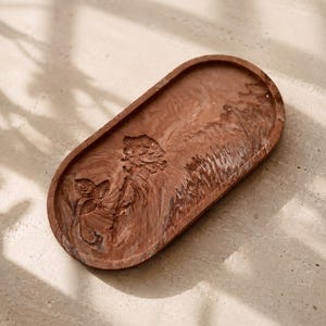 K&ouml;nnte beinhalten: Eine braune, ovale dekorative Schale mit einem geschnitzten Blumendesign. Die Schale hat einen erh&ouml;hten Rand und ein glattes Inneres. Das Blumendesign umfasst eine Rose und einen Vogel. Die Schale besteht aus einem festen Material und eignet sich f&uuml;r kleine Gegenst&auml;nde.