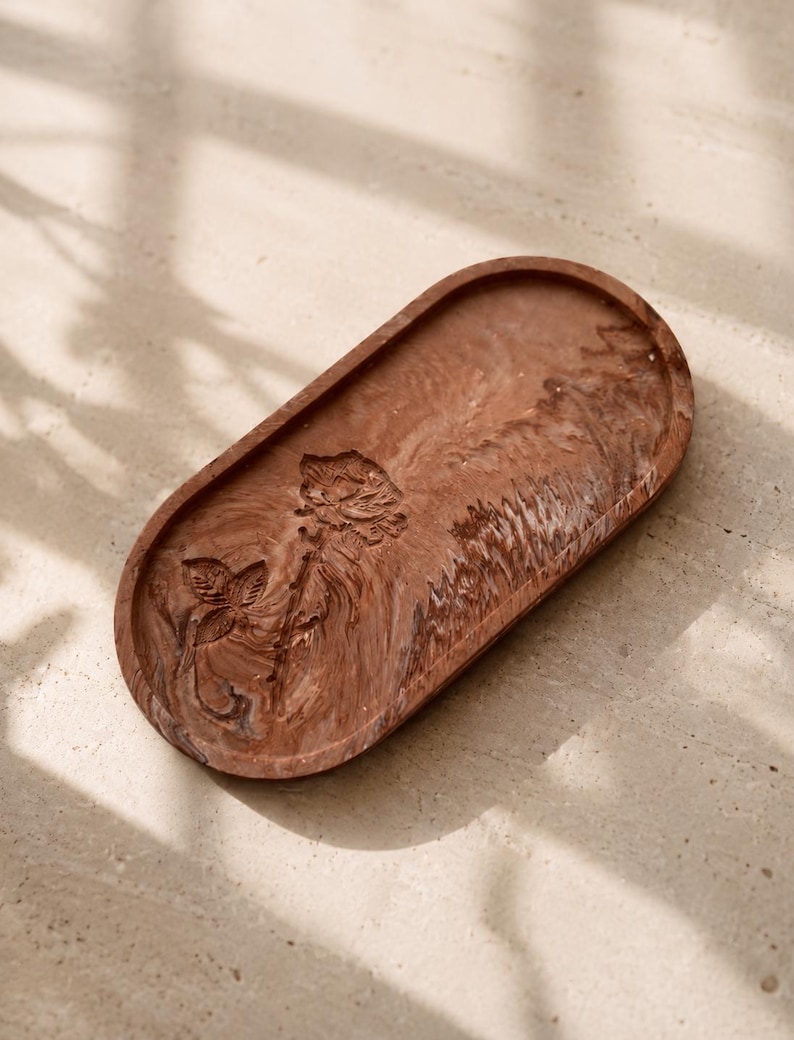 K&ouml;nnte beinhalten: Eine braune, ovale dekorative Schale mit einem geschnitzten Blumendesign. Die Schale hat einen erh&ouml;hten Rand und ein glattes Inneres. Das Blumendesign umfasst eine Rose und einen Vogel. Die Schale besteht aus einem festen Material und eignet sich f&uuml;r kleine Gegenst&auml;nde.