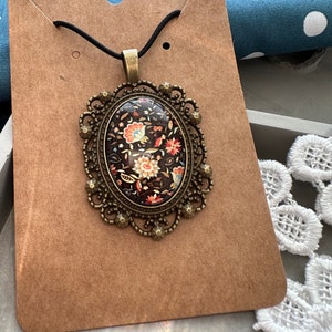 May include: A vintage-style pendant necklace featuring a black and brown cameo with a floral design. The cameo is set in a gold-toned metal frame with intricate details.