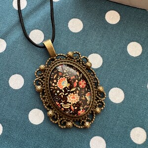 May include: A black and gold oval pendant necklace with a floral pattern inside the glass. The necklace is on a blue and white polka dot fabric background.