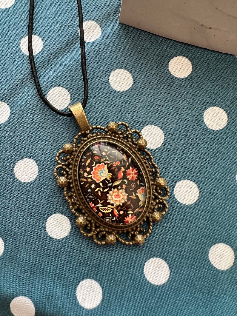 May include: A black and gold oval pendant necklace with a floral pattern inside the glass. The necklace is on a blue and white polka dot fabric background.
