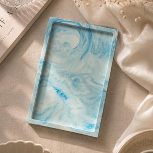 Op de afbeelding: Een rechthoekige dienblad met een gemarmerd blauw en wit ontwerp. De dienblad heeft een opstaande rand en een glad oppervlak. De dienblad staat op een neutraal gekleurd oppervlak. Decoratieve items staan op de achtergrond.
