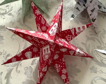 Puede incluir: Una colección de adornos de estrellas de papel en varios colores y patrones. La estrella central es roja con un diseño blanco de temática navideña. Otras estrellas son verdes y blancas, con patrones de árboles y estrellas. Estas estrellas decorativas son ideales para la decoración navideña.
