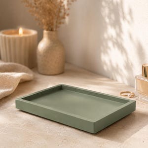 May include: A rectangular, sage green tray. The tray is made of a solid material and has a slightly raised edge. The tray is empty and sits on a neutral-toned surface. A lit candle, a small vase with dried flowers, and a perfume bottle are in the background.