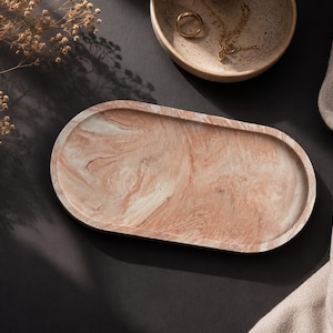 K&ouml;nnte beinhalten: Ein ovales dekoratives Tablett mit einem marmorierten rosa-wei&szlig;en Muster. Das Tablett steht auf einer dunklen Oberfl&auml;che, mit einer kleinen Schale und Stoff im Hintergrund. Eine goldene Halskette und ein Ring befinden sich in der Schale.