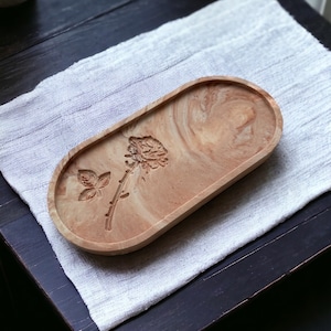 K&ouml;nnte beinhalten: Ein ovales Tablett aus Harz mit Marmoreffekt in Beige- und Braunt&ouml;nen. Das Tablett zeigt ein gepr&auml;gtes Rosen- und Schmetterlingsmotiv. Es liegt auf einem wei&szlig;en, strukturierten Tuch, wahrscheinlich f&uuml;r dekorative oder praktische Zwecke im Haushalt bestimmt.