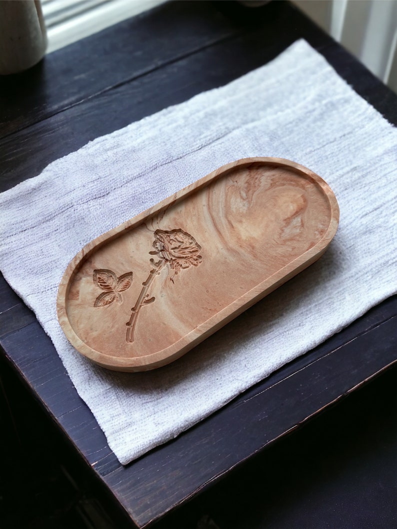 K&ouml;nnte beinhalten: Ein ovales Tablett aus Harz mit Marmoreffekt in Beige- und Braunt&ouml;nen. Das Tablett zeigt ein gepr&auml;gtes Rosen- und Schmetterlingsmotiv. Es liegt auf einem wei&szlig;en, strukturierten Tuch, wahrscheinlich f&uuml;r dekorative oder praktische Zwecke im Haushalt bestimmt.