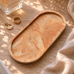 K&ouml;nnte beinhalten: Ein ovales Holztablett mit einem marmorierten Muster in Beige- und Braunt&ouml;nen. Ein goldener Ring und eine Halskette mit Anh&auml;nger liegen neben dem Tablett. Ein Parf&uuml;mflakon und ein Buch sind ebenfalls im Bild.