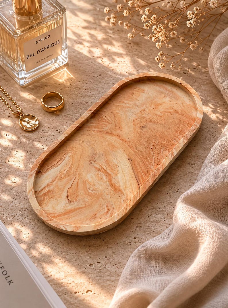 K&ouml;nnte beinhalten: Ein ovales Holztablett mit einem marmorierten Muster in Beige- und Braunt&ouml;nen. Ein goldener Ring und eine Halskette mit Anh&auml;nger liegen neben dem Tablett. Ein Parf&uuml;mflakon und ein Buch sind ebenfalls im Bild.
