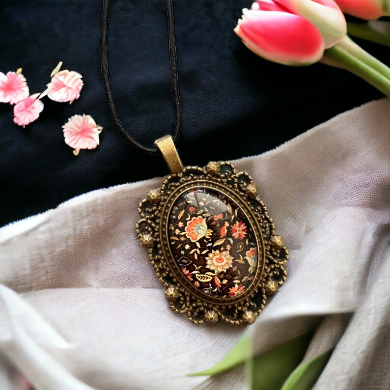 May include: A gold-toned pendant necklace with a black and white floral design inside a clear oval cameo. The necklace is on a black fabric background.