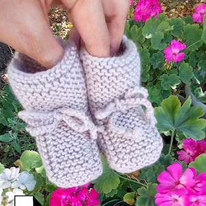May include: A pair of light beige knitted baby booties with delicate ties. The booties are held up against a backdrop of vibrant pink flowers and green foliage, showcasing a handmade craft item.