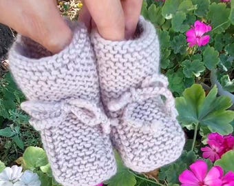 Op de afbeelding: Een paar lichtbeige gebreide babyslofjes met delicate strikjes. De slofjes worden omhoog gehouden tegen een achtergrond van levendige roze bloemen en groen gebladerte, wat een handgemaakt ambachtelijk item laat zien.