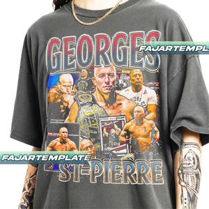 May include: Dark grey t-shirt featuring a collage of images of Georges St-Pierre, with the name "GEORGES ST-PIERRE" in a distressed font. The shirt has a vintage look, with a gold chain necklace.
