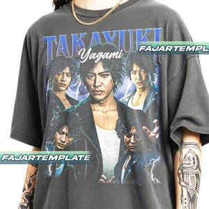 以下が含まれることがあります： 高木由貴のグラフィックと青い文字が特徴の濃いグレーのTシャツ。デザインには人物の複数の画像が含まれています。シャツは半袖でゆったりとしたフィット感です。「FAJARTEMPLATE」の文字も表示されています。
