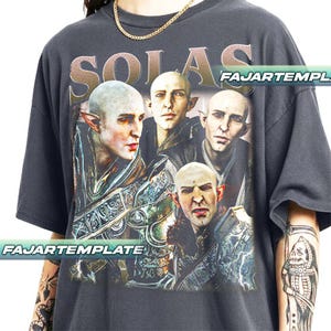 Peut inclure: T-shirt gris foncé avec un graphisme de quatre personnages aux oreilles pointues et chauves. Le mot "SOLAS" est imprimé au-dessus du graphisme en couleur or. Le t-shirt porte l'inscription "FAJARTEMPLE".