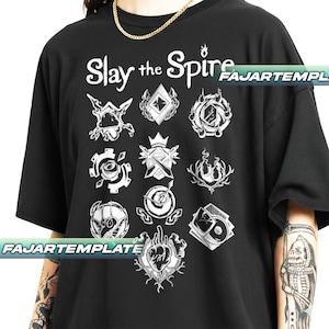 May include: Black t-shirt with the text "Slay the Spire" and various white graphic designs. The designs include geometric shapes, gears, and other stylized emblems. The shirt has a crew neck and short sleeves.