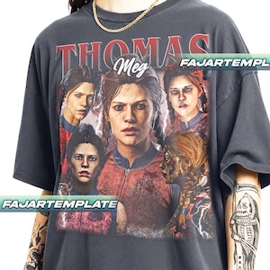 May include: Dark gray t-shirt featuring a graphic of a woman with the text "THOMAS Meg" in red. The graphic includes multiple images of the woman with lightning bolt accents. The text "FAJARTEMPLE" is printed on the bottom.