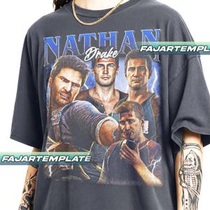 Może przedstawiać: Ciemnoszara koszulka z grafiką Nathana Drake'a i napisem "NATHAN Drake" w kolorze niebieskim i białym. Projekt zawiera wiele zdjęć postaci z błyskawicami. Koszulka ma wygląd vintage.