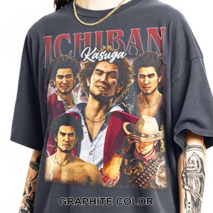 Peut inclure: Un t-shirt gris anthracite avec une impression graphique d'Ichiban Kasuga dans diverses poses et tenues. Le texte ICHIBAN apparaît en grandes lettres rouges en haut, avec Kasuga écrit en écriture blanche en dessous. GRAPHITE COLOR est en bas.