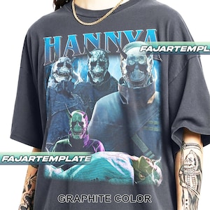 Può includere: T-shirt color grafite con una grafica che presenta la parola "HANNYA" in blu. La grafica raffigura più figure con maschere a forma di teschio e il testo "FAJARTEMPLATE" in bianco. La maglietta ha maniche corte.