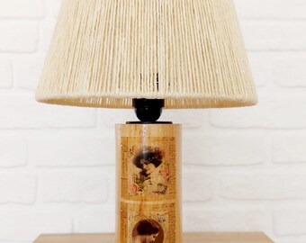 May include: A table lamp with a cream-coloured, rope-wrapped lampshade and a cylindrical wooden base. The base is decorated with three vintage images of women.