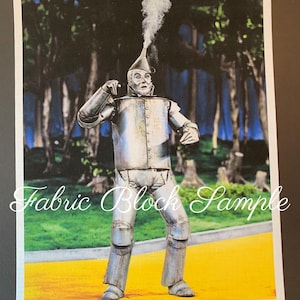 May include: A fabric block sample featuring a printed image of the Tin Man character from The Wizard of Oz. The figure is metallic silver, wearing a funnel hat with smoke rising from the top, standing on a yellow path in a forest setting.