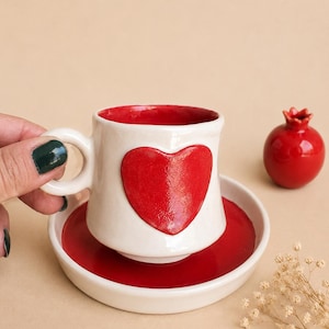Puede incluir: Un juego de taza y platillo de cerámica con un exterior blanco brillante y un gran corazón rojo en relieve en la parte frontal. El interior de la taza y el centro del platillo tienen un acabado en esmalte rojo vibrante, junto a una granada roja.
