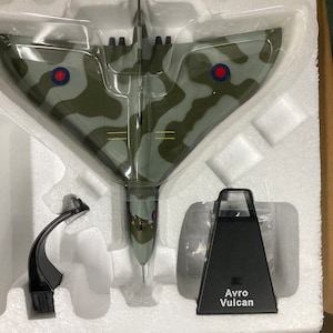 May include: A model of a Avro Vulcan bomber in a camouflage paint scheme with red roundels. The model is displayed on a white foam base with a black stand. The stand has the text "Avro Vulcan" printed on it.