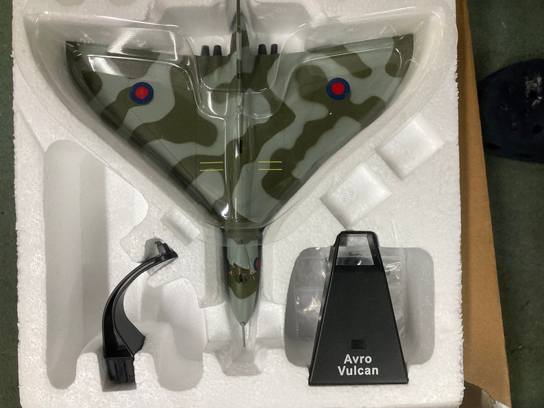 May include: A model of a Avro Vulcan bomber in a camouflage paint scheme with red roundels. The model is displayed on a white foam base with a black stand. The stand has the text "Avro Vulcan" printed on it.