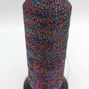 Könnte beinhalten: Eine Spule mit mehrfarbigem Metallic-Garn mit schwarzem Sockel. Das Garn ist eine Mischung aus Schwarz, Gold, Blau und Rosa.